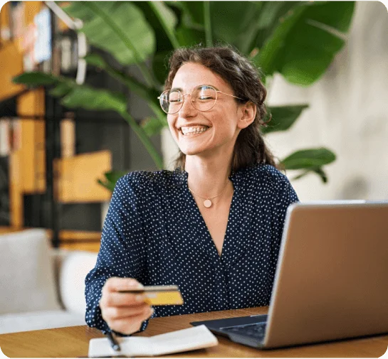 A smiling woman sitting at a desk, holding a bank card and using a laptop.