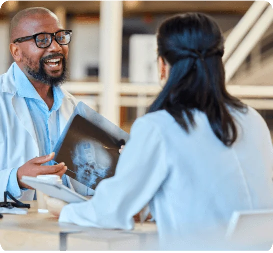 A male doctor smiling and talking to a female doctor represents a clinical consultation. The male doctor is holding an X-ray of a person.